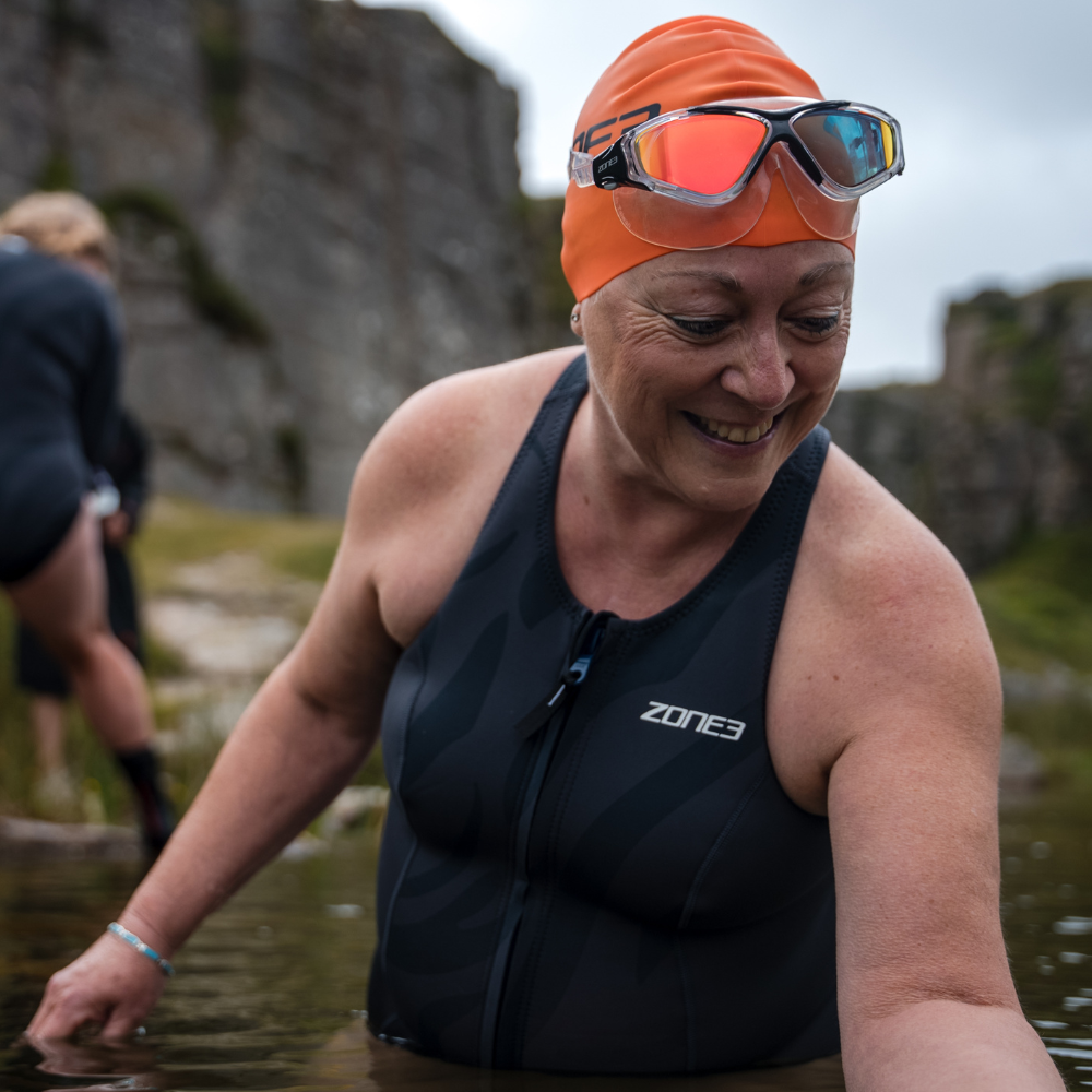 Lächelnde Schwimmerin mit weißer Zone3-Badekappe und Vision Max-Schwimmmaske auf der Stirn, teilweise im Wasser im Freien eingetaucht.