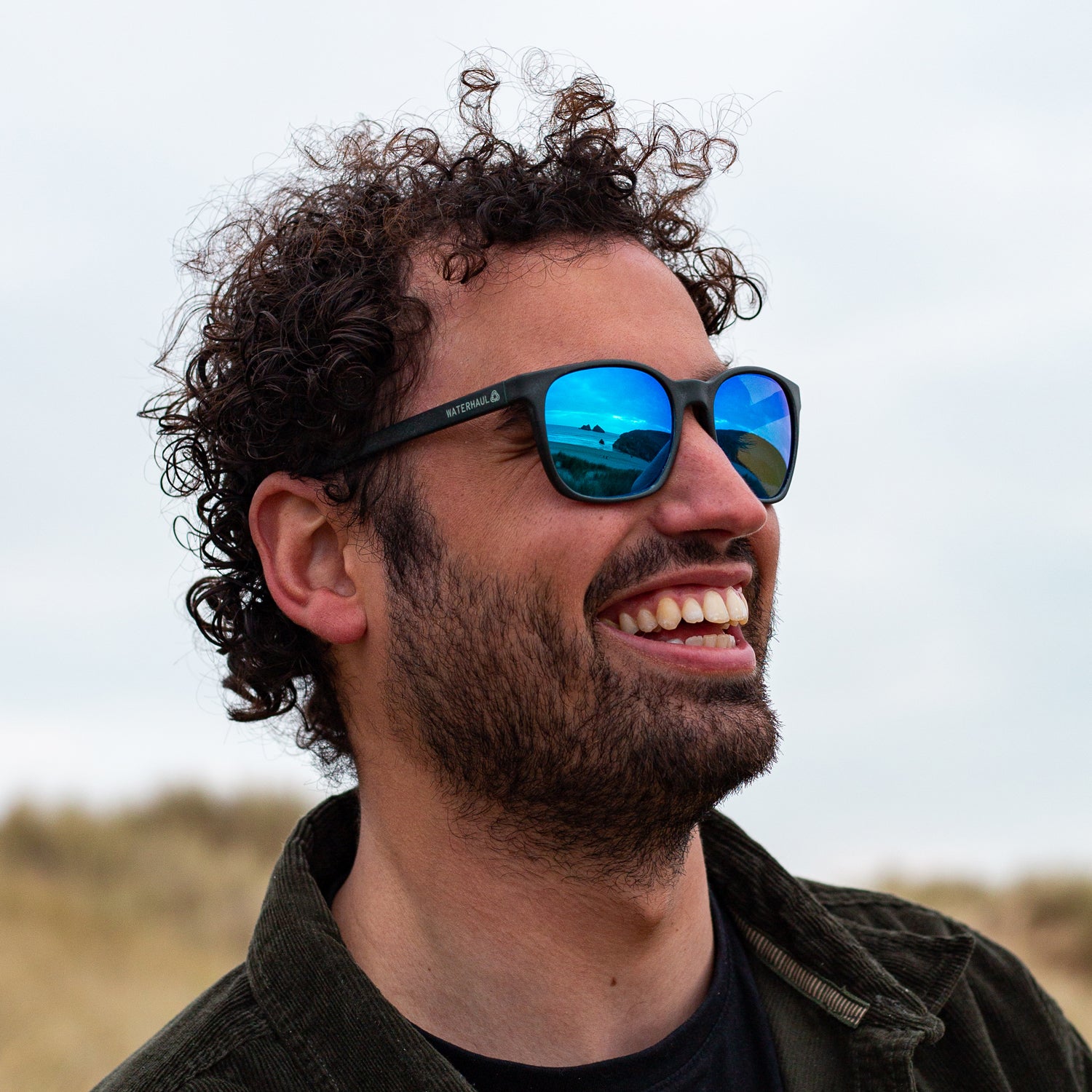 Smiling man wearing Waterhaul sunglasses with matte dark grey frames and blue mirrored polarized lenses, reflecting a coastal landscape. The image captures an outdoor, eco-conscious vibe with natural background and relaxed energy.