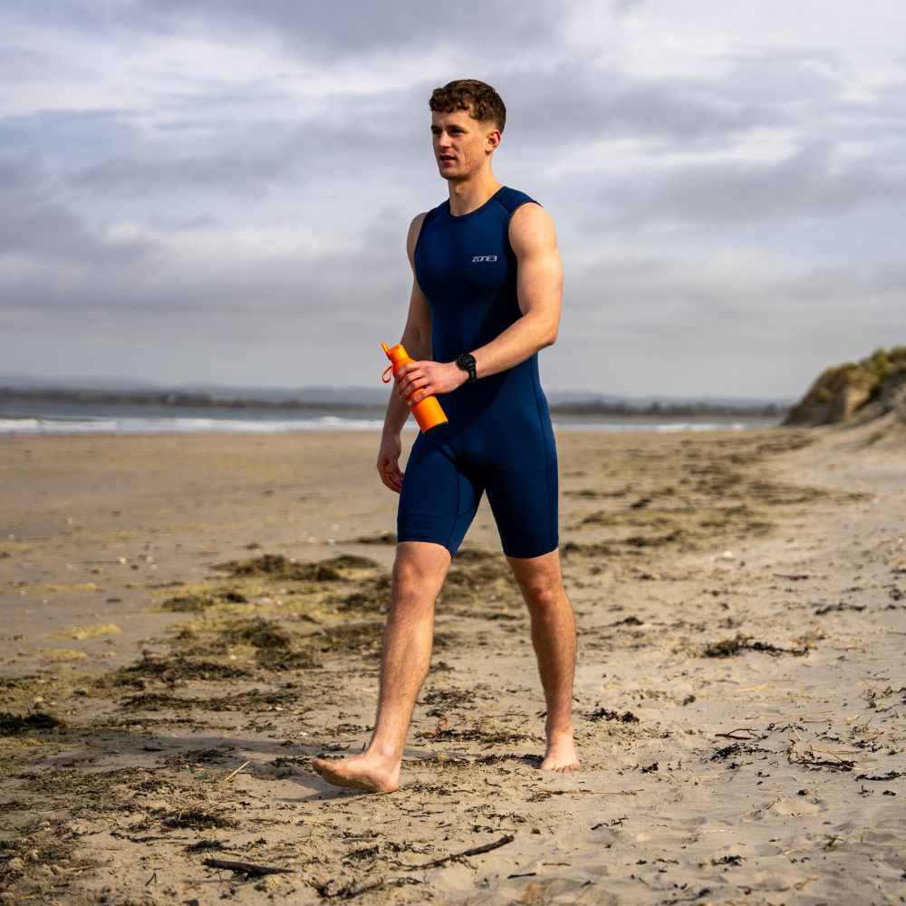 Person wearing blue Yulex kneeskin wetsuit walks on a beach holding an orange water bottle