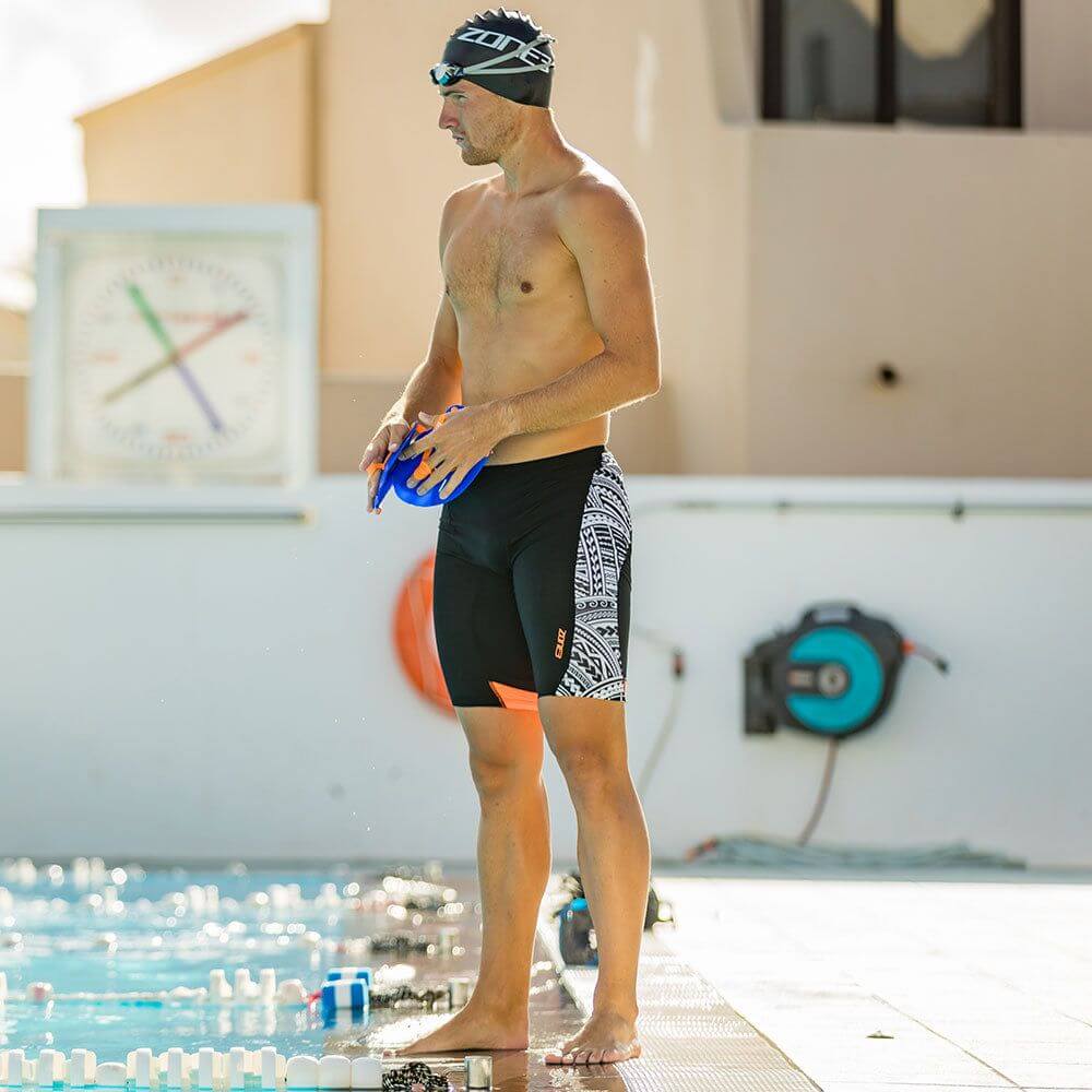 Man standing poolside wearing black swim jammers with white Kona Speed print on the side and orange accents.