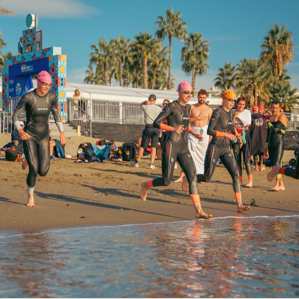 Athletes wearing ZONE3 wetsuits running into the sea for their swim start. In the background there are palm trees and spectators.